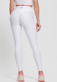 Žena v texturovaných bílých legínách s vysokým pasem a odpovídajícím crop topem, doplněná bílými teniskami s růžovými akcenty, zobrazená zezadu.