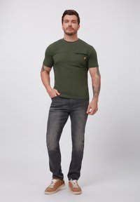 Oliven grøn kortærmet t-shirt med brystlomme og et gult logo. Sammen med mørkegrå jeans og lysebrune sneakers.