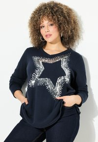 Marineblauer Strickpullover mit einem großen silbernen Sterndesign aus Pailletten. Lange Ärmel, lockere Passform und Taschen-Detail.