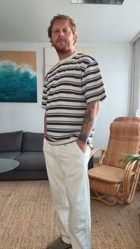 T-shirt in cotone a righe nei colori beige, marrone e blu; abbinato a pantaloni bianchi a vestibilità dritta. Mostra un tatuaggio sul braccio sinistro.