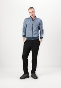 Jachetă albastră cu model, cu fermoar, având dungi albe pe mâneci, asociată cu pantaloni negri. Modelul stă cu mâinile în buzunare, purtând teniși negri.