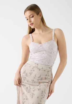Femme portant une camisole en dentelle rose clair et une jupe beige avec broderie florale, posant devant un fond blanc uni.