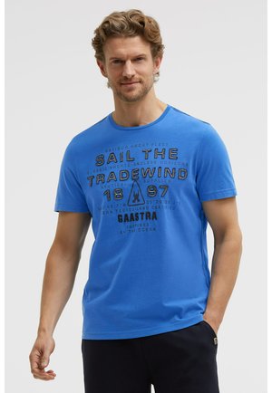Mann mit lockigem Haar, der ein blaues T-Shirt mit schwarzem nautischem Text trägt, steht mit einer Hand in der Tasche vor einem schlichten Hintergrund.