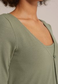 Ribbade, olivgröna koftor med en djup V-ringning, med knäppning och en strukturerad tyg. Slät, figurnära design som framhäver formen.