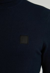 Marineblå stickad tröja med hög krage. Har en fyrkantig svart logotyp på vänster bröst och räfflade detaljer vid halsen och axlarna.