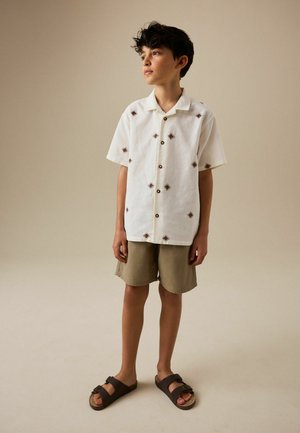 Junge mit lockigem dunklem Haar, der ein weißes kurzärmeliges Hemd mit braunen Rautenmuster, beige Shorts und dunkelbraune Sandalen trägt und vor einem schlichten Hintergrund steht.