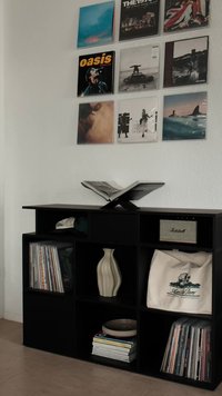 Juodas lentynos vienetas su kvadratinėmis ertmėmis. Viršuje yra smėlio spalvos vaza, audinio maišelis, vinilo plokštės ir atvira knygų stovė.