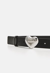 Zwarte leren riem met een glanzende zilveren hartvormige gesp met reliëflettering "HUGO." Verstelbaar met meerdere gaatjes voor maat.