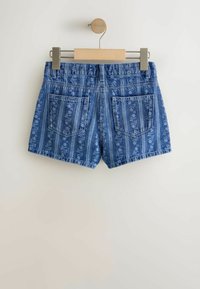 Denim lühikesed püksid lillemustriga, sinistes toonides, kahe tagataskuga. Omavad õmblemata äärt ja standardset vöökorda. Riputatud puidust ja metallist riidepuule.