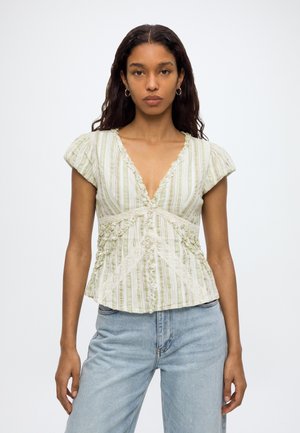 Jonge vrouw met lang donker haar, die een lichtgroene en wit gestreepte blouse met kanten details en lichtblauwe spijkerbroek draagt, staand tegen een witte achtergrond.