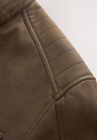 Close-up van de schouder van een bruine leren jas met gestikte doorgestikte details en naadlijnen tegen een neutrale achtergrond.