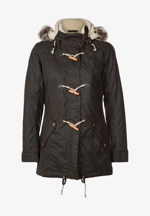 Woman wearing a dark green hooded parka with fur trim, white knit sweater, gray skinny jeans, and black ankle boots standing with one hand in pocket.