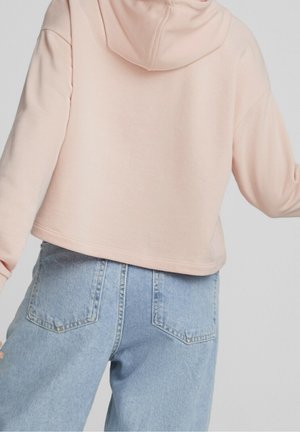 Persona con una sudadera corta de color rosa pálido y vaqueros de cintura alta de color azul claro, vista desde atrás sobre un fondo liso.