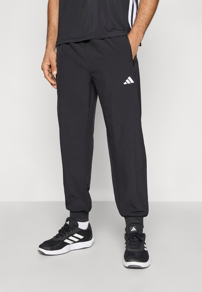 Sorte atletik-joggers med elastiske manchetter, sidelommer og hvidt Adidas-logo. Båret med sorte atletiksko med hvide striber.