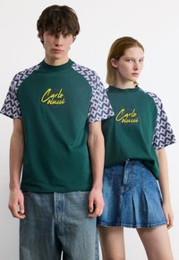 Νέος άνδρας και γυναίκα φορούν ταιριαστά πράσινα πουκάμισα με μοτίβο στα μανίκια και λογότυπο "Carlo Colucci", συνδυασμένα με τζιν παντελόνι και φούστα.