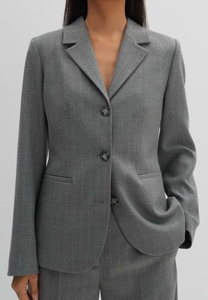 Femme portant un blazer gris à fines rayures ajusté avec trois boutons, assorti à un pantalon coordonné, une main dans la poche, fond neutre.