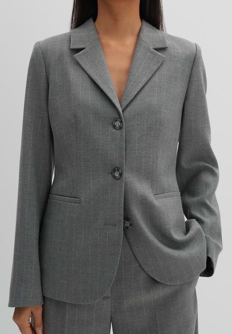Femme portant un blazer gris à fines rayures ajusté avec trois boutons, assorti à un pantalon coordonné, une main dans la poche, fond neutre.