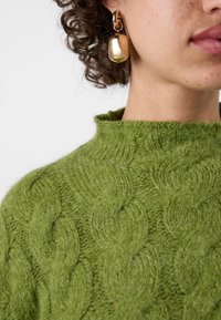Pull vert en tricot torsadé avec un col rétracté, associé à une boucle d'oreille en or brillant avec un accent lisse et arrondi.