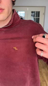 Person wearing a maroon quarter-zip sweater with a small embroidered moose on the chest, pointing to the emblem with their right hand.