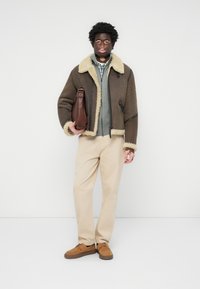 Chaqueta de borrego marrón con forro beige, camisa gris con cremallera, pantalones beige y zapatos de ante marrón, sosteniendo un bolso de cuero marrón.