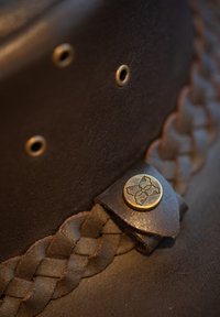 Brown leather hat with braided band and gold metal accent featuring a floral design. Textured surface with stitching details.