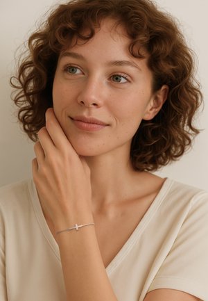 Silverarmband med ett korscharm hängande på ett tunt kedja, buren på en kvinnas handled. Armbandet har en slät yta och en minimalistisk design.