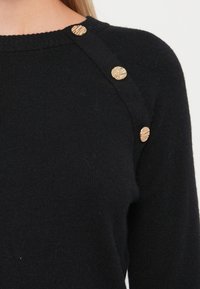 Pull noir en tricot avec un col rond, présentant des boutons dorés texturés le long de l'épaule gauche pour une touche de détail supplémentaire.