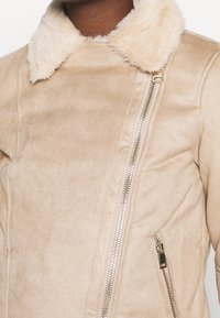 Chaqueta de ante sintético beige con un suave collar de piel. Presenta un cierre diagonal y bolsillos laterales con acentos en negro contrastantes.