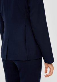 Marinblå kavaj med skräddarsydd passform, slät textur och diskret ryggslitsdetalj. Kombinerad med matchande byxor som visar en enhetlig design.