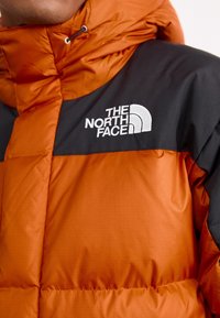 Doudoune avec un corps orange et des panneaux d'épaule noirs, avec une texture matelassée et un logo blanc sur la partie gauche de la poitrine.