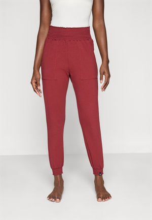 Femeie îmbrăcată cu pantaloni jogger burgundy, cu talie înaltă, bandă elastică la talie și la pulpe, stând desculță pe un fundal simplu.