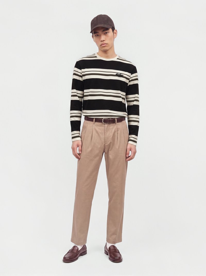 Un mannequin se tient devant un fond blanc, vêtu d’une casquette foncée, d’un pull à rayures noires et crème, d’un pantalon beige et de mocassins bordeaux.