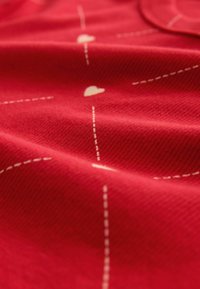 Roter Stoff mit kleinen weißen Herzmustern und gestrichelten Linien, wobei die gerippte Textur im gesamten Material sichtbar ist.