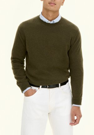 Homme portant un pull vert olive par-dessus une chemise bleu clair à col, pantalon blanc et ceinture noire, main dans la poche sur fond uni.
