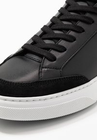 Svart läder- och mockasneaker med platta svarta snören, en strukturerad vit gummisula och sydda detaljer längs de övre kanterna.