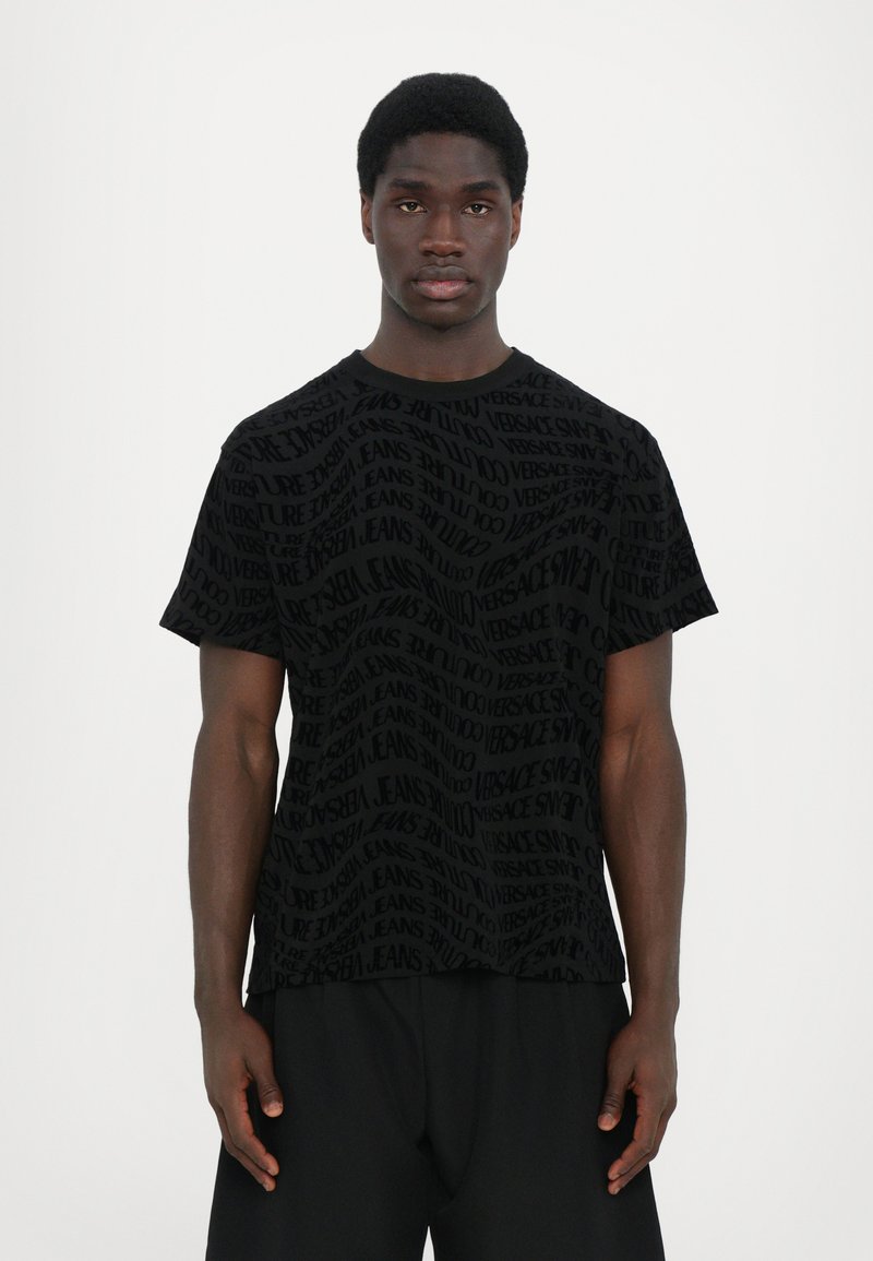 Juodas trumpas rankovės T-shirt, kuris turi tekstūruotą raštą su žodžiu "Versace" įvairiomis orientacijomis. Audinys atrodo lygus ir lengvas.