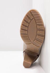 Semelle de botte beige avec des motifs de bande de roulement texturés et un talon bloc, positionnée verticalement sur fond blanc.