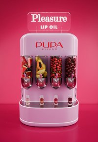 Dozownik olejku do ust w kolorze różowym z przezroczystymi pojemnikami zawierającymi arbuz, papaję, truskawki i wiśnie; poniżej wyświetlane są małe buteleczki z olejkiem do ust.