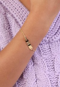 Gouden armband met een vlak, rechthoekig middengedeelte en een fijne ketting, gedragen om een pols tegen een gestructureerde lavendelkleurige gebreide achtergrond.