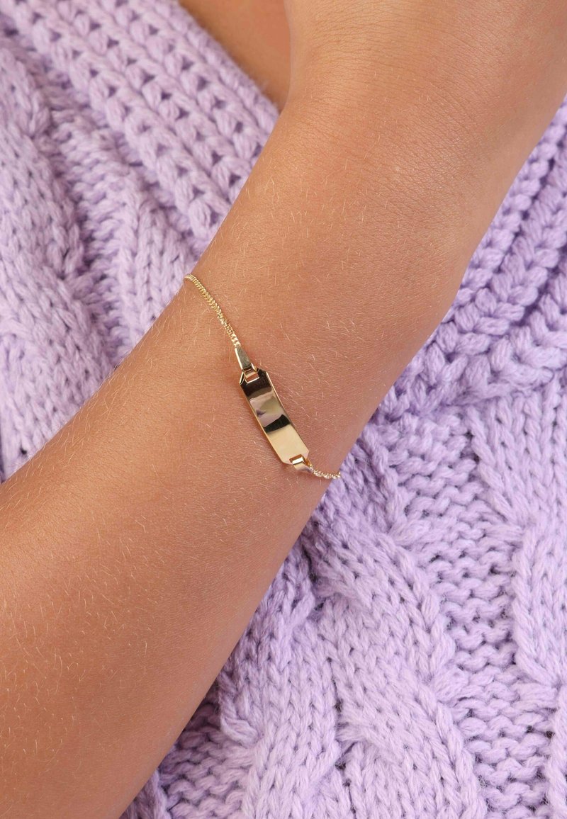 Gouden armband met een vlak, rechthoekig middengedeelte en een fijne ketting, gedragen om een pols tegen een gestructureerde lavendelkleurige gebreide achtergrond.