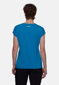 Blå t-shirt med kort ärm, rundad halsringning och uppvikta ärmar. Har en vit logotyp på baksidan nära halsen. Slät textur.