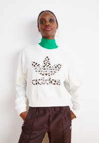 Hvit sweatshirt med leopardmønster og Adidas-logo. Har rund hals og avslappet passform. Grønn turtleneck under, brune mønstrede bukser.