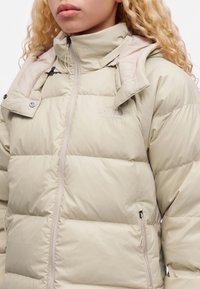 Μπεζ μπουφάν North Face με επένδυση, ψηλό γιακά, φερμουάρ μπροστά και φερμουάρ στο πλαϊνό τσέπη, φορεμένο από άτομο με ξανθά σγουρά μαλλιά.