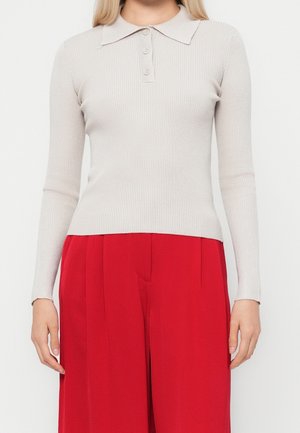 Femme portant un polo côtelé beige clair à manches longues associé à un pantalon large taille haute rouge vif.