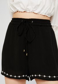 Svarta vävda shorts med elastisk midja, dragsnören och vita broderade detaljer längs fållen. Mjuk textur, lös passform.
