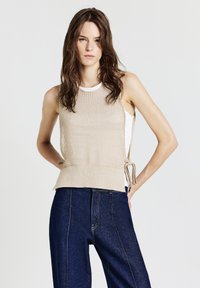 Donna con lunghi capelli castani indossa un top a maglia beige senza maniche sopra una camicia bianca e jeans a vita alta blu scuro, in piedi con le mani sui fianchi.