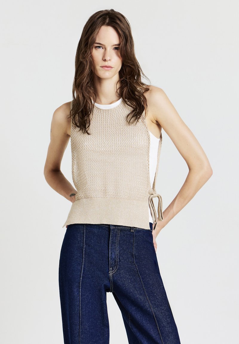 Donna con lunghi capelli castani indossa un top a maglia beige senza maniche sopra una camicia bianca e jeans a vita alta blu scuro, in piedi con le mani sui fianchi.