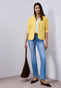 Femme debout portant un blazer jaune, une chemise blanche, un jean bleu, une ceinture noire et des chaussures noires, tenant un grand sac à main marron foncé.