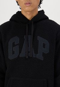 Felpa con cappuccio nera in pile con superficie texturizzata, caratterizzata da un logo "GAP" nero sul davanti, con cordini e tasca frontale.