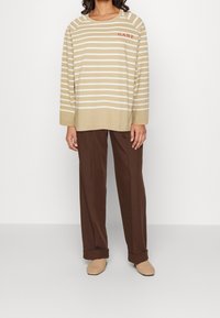 Beige gestreiftes Langarmoberteil mit rotem "GANT"-Logo, kombiniert mit braunen schmal zulaufenden Hosen und hellen, gewebten Slippern. Glatte Textur.